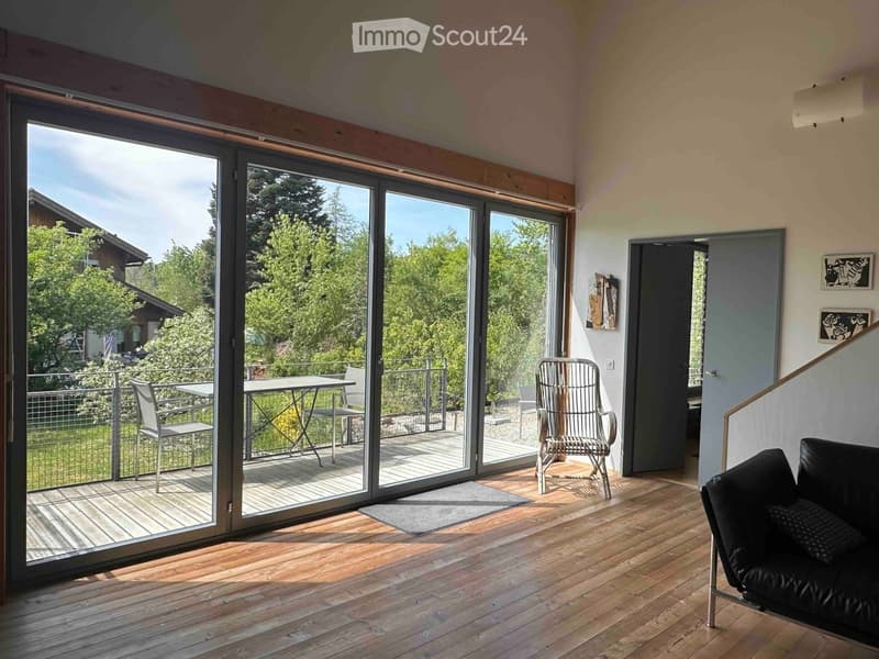 Bright living room with wooden flooring, large sliding glass doors opening to a balcony, a black leather sofa, a wicker chair, and a small table on the balcony.