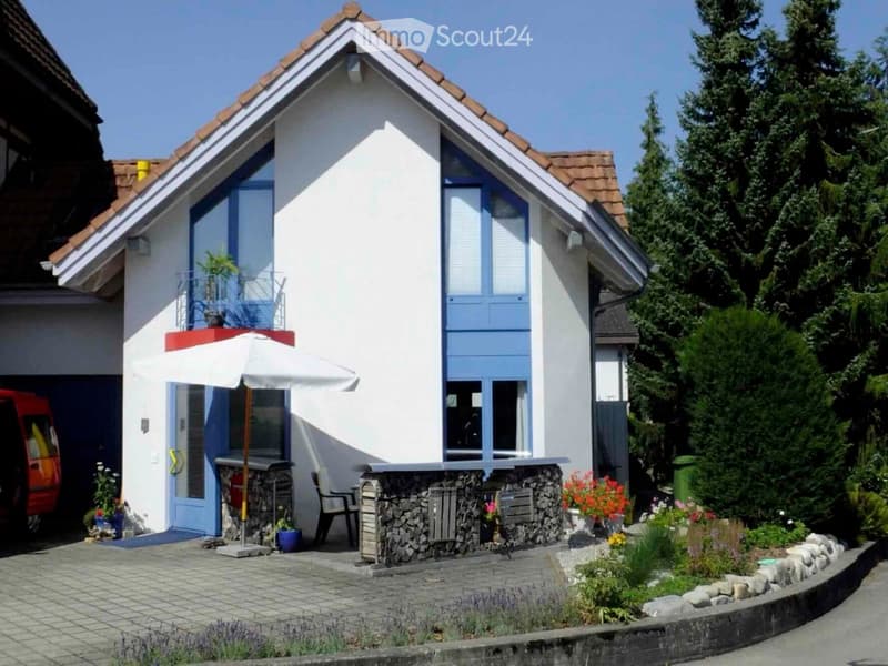 Single-story house with white walls, blue shutters, and a red-tiled roof. Features a small balcony with plants, a patio area with a table, chairs, and an umbrella, surrounded by a well-maintained garden with flowers and shrubs.