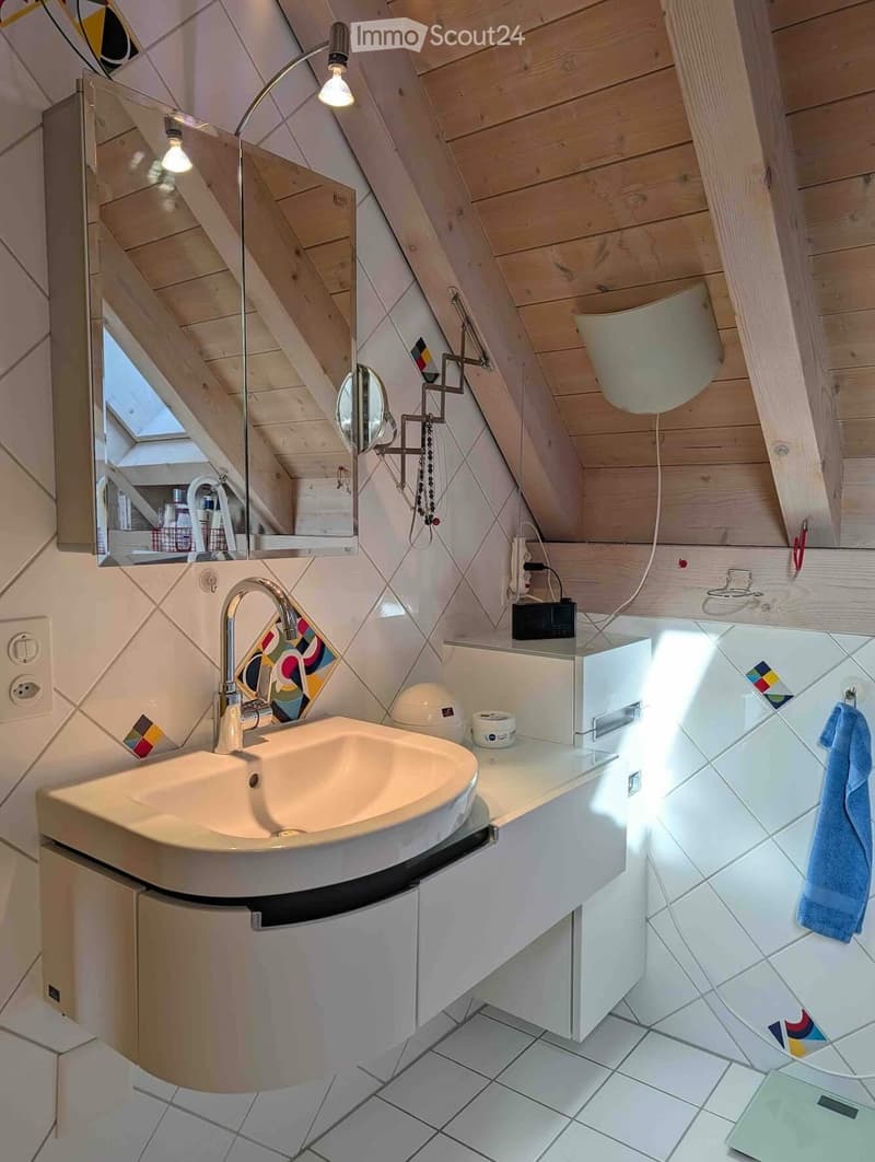 Modern bathroom with a white sink, mirror, and wooden ceiling beams. Features include a towel, light fixture, and decorative tiles.