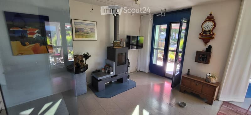 Modern living room with a fireplace, television, and large windows. Features include a wooden table, chairs, picture frames, a decorative clock, and a small plant.
