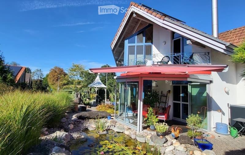 Modern two-story house with a red roof, large windows, and a spacious garden area featuring a pond, plants, and outdoor furniture.