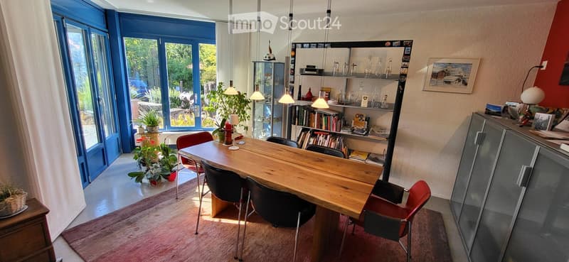 Bright, modern dining area with a wooden table, chairs, bookshelf, and decorative plants. Large windows provide natural light and a view of the outdoor area.