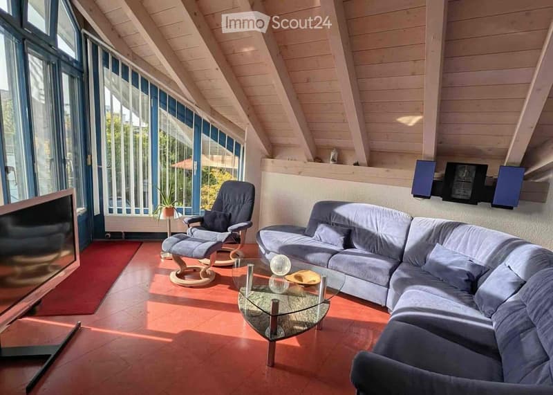 Modern living room with a grey sectional sofa, a single armchair, a glass coffee table, and a flat-screen TV. The room features wooden beams on the ceiling and large windows with blue frames, allowing ample natural light.