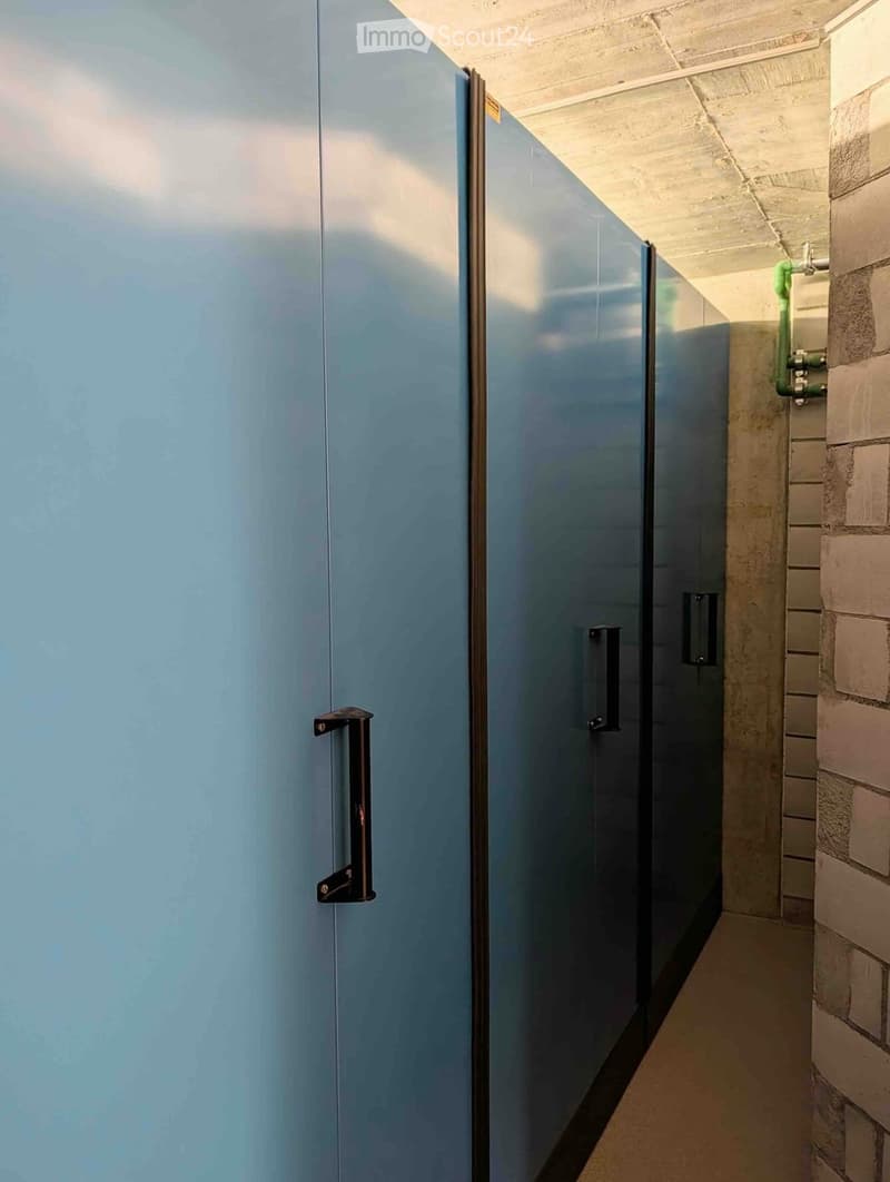 A narrow hallway with light blue walls, a polished concrete floor, and a closed door with a metallic handle on the left side. Exposed brickwork is visible on the right wall, along with some pipes and fixtures.
