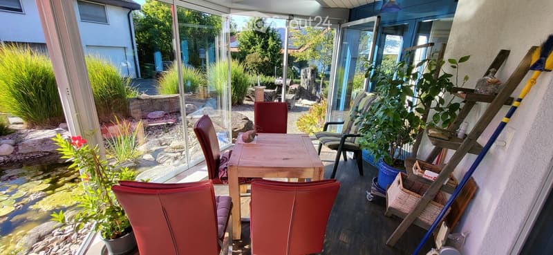 Bright, enclosed balcony with wooden table, red chairs, and potted plants, overlooking a landscaped garden with a pond.