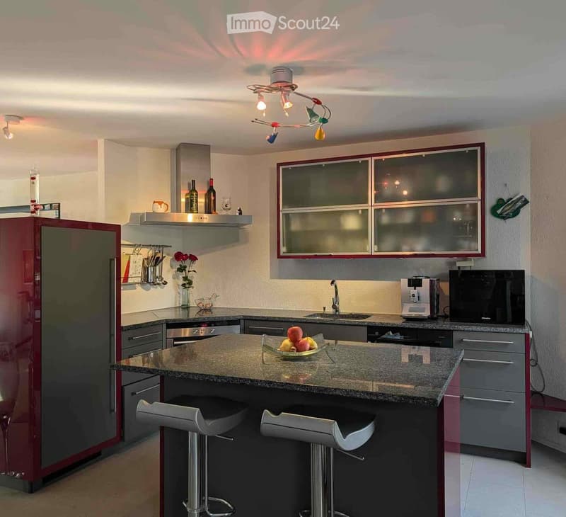 Modern kitchen with granite countertop, red refrigerator, built-in oven, microwave, coffee maker, and two stools at the island.