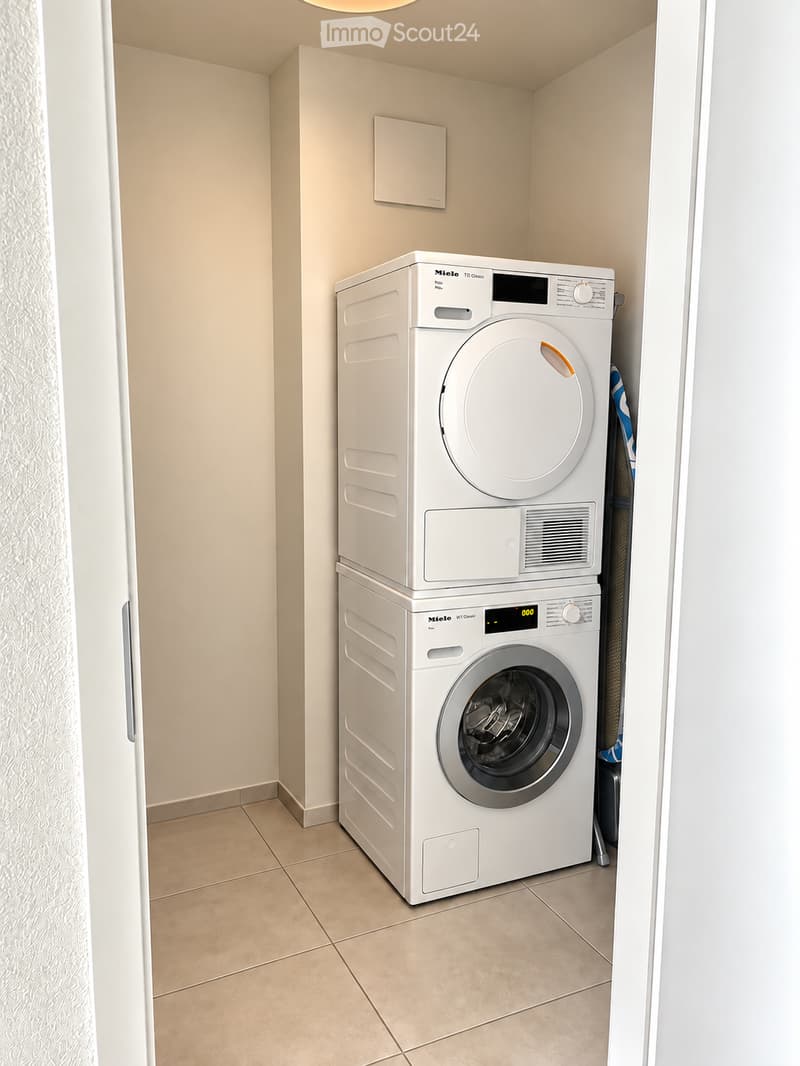 Compact bathroom with a stacked washing machine and dryer unit, beige tiled floor, and light-colored walls.