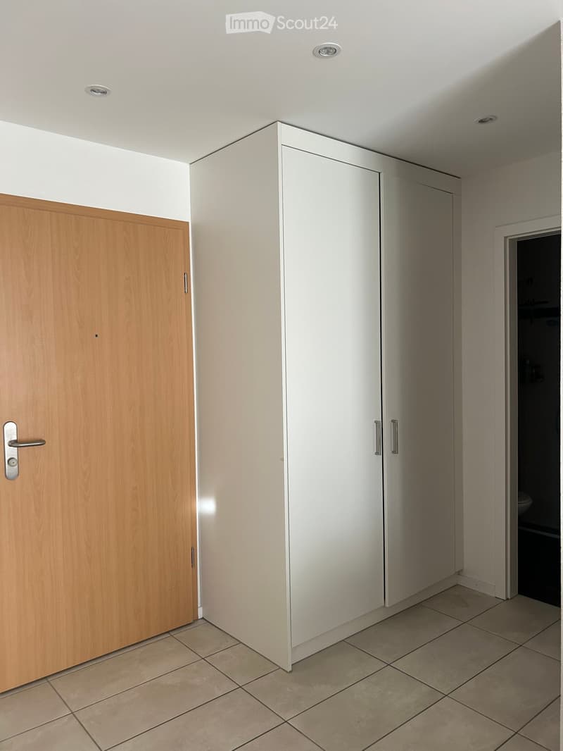Empty hallway with tiled floor, white walls, and built-in white cabinets. A wooden door is visible on the left, and a doorway leads to another room on the right.