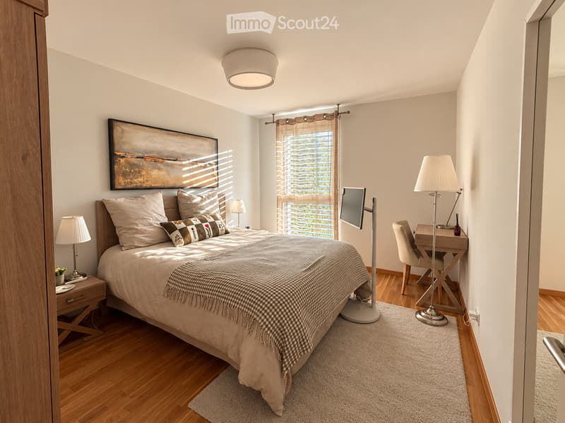 Modern bedroom with a large bed, nightstand, desk, and a large window with blinds. The room features a cozy bed with pillows and a blanket, a small nightstand with a lamp, a desk with a chair, and a mounted screen on the wall.