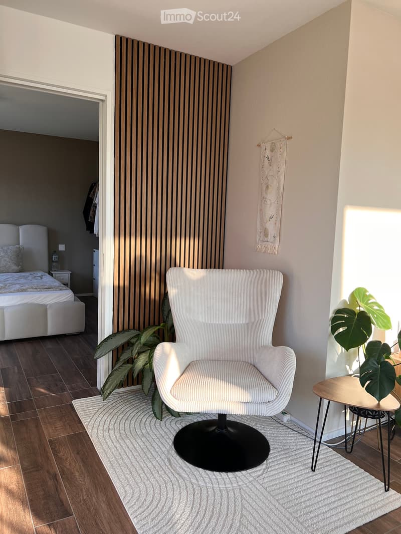 A bedroom with a bed, wooden floors, wall-mounted artwork, a potted plant, a cozy armchair, and a white rug.