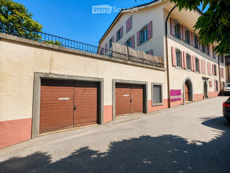 Two-story building with multiple garage doors, pink shutters, and a raised ground floor with a balcony.