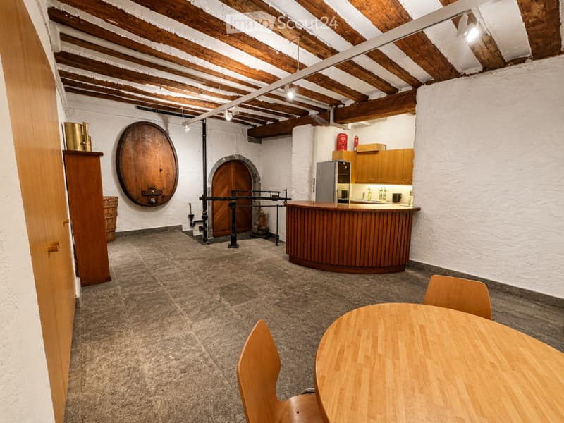 The room features exposed wooden beams on the ceiling, a round wooden table with chairs, a bar counter with a refrigerator, and a large wooden wheel on the wall.