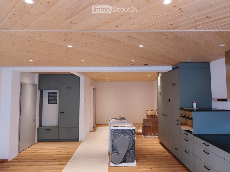 Modern kitchen with grey cabinets, wooden flooring, and exposed wooden ceiling beams. A dishwasher is visible on the floor, and a refrigerator is partially visible in the background.