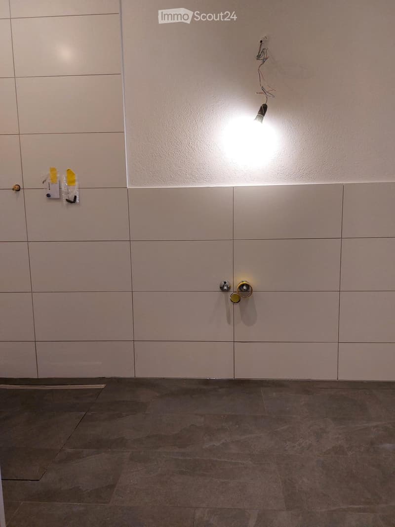 Empty room with beige tiled walls and a dark tiled floor. A light fixture is attached to the wall, and there are some small items and cables visible on the left side.