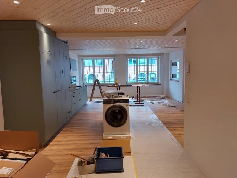 Empty room with wooden flooring, large windows, and a private washing machine placed in the center. The room is under renovation with construction materials and tools visible.