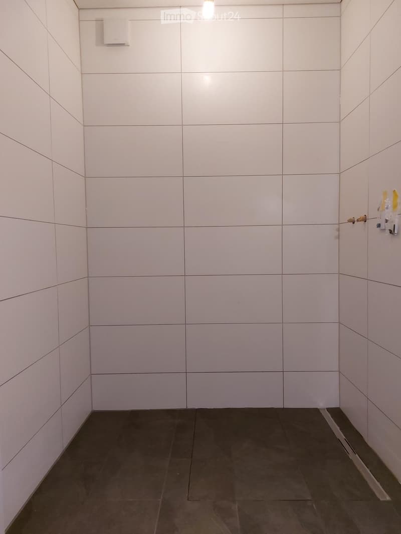 Empty room with white tiled walls and dark tiled floor, featuring a light fixture and a small shelf on the wall.
