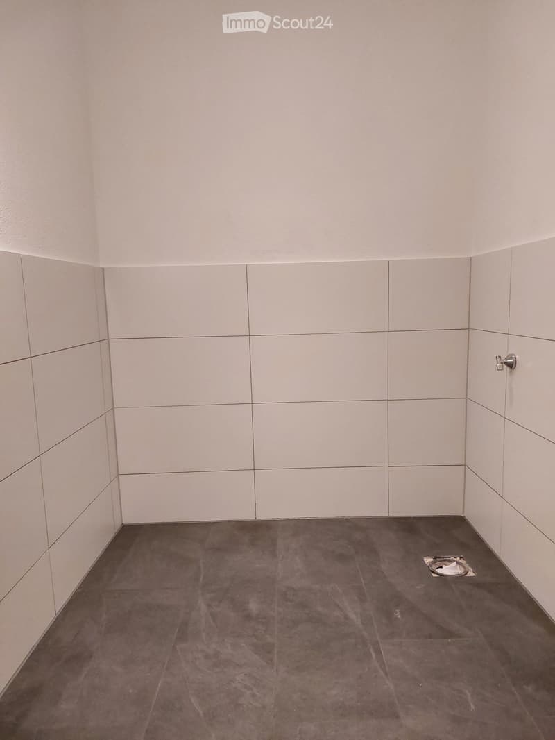 Empty room with white tiled walls and a dark grey concrete floor, featuring a small drain in the corner.
