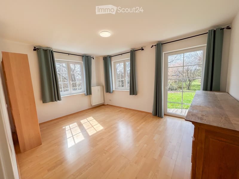 Empty room with wooden flooring, large windows with green curtains, and a radiator. Natural light streams in from the windows.