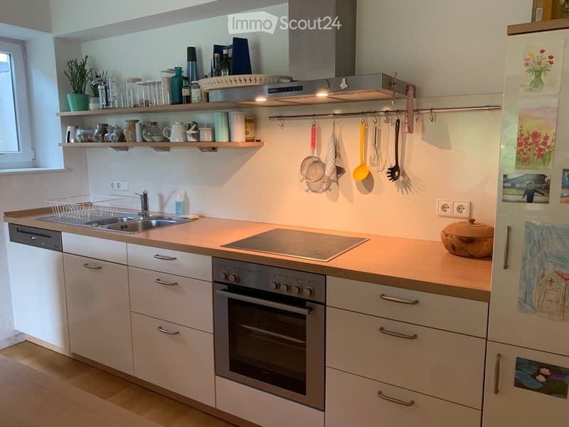 Modern kitchen with white cabinets, light wooden countertops, stainless steel appliances, and open shelving with jars, bottles, and decorative items.