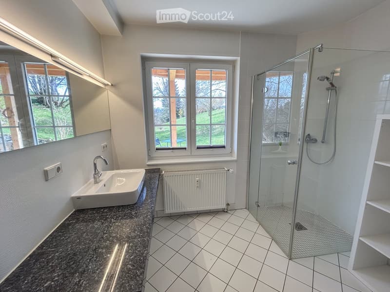 Modern bathroom with a white tiled floor, granite countertop, sink, shower with glass enclosure, radiator, and large windows offering a view of the garden.