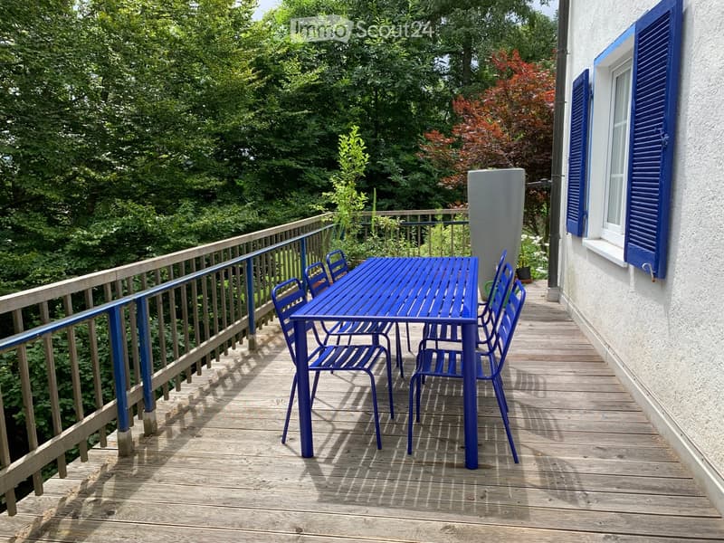 Balcony with wooden decking, blue metal table and chairs, railing, and lush greenery in the background.