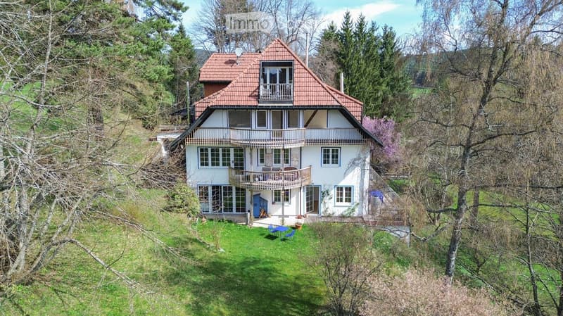 Two-story house with red-tiled roof, white walls, multiple windows, balconies, and a small garden area with outdoor furniture.