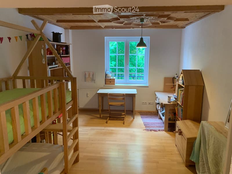 Cozy bedroom with wooden bunk bed, desk, chair, bookshelf, radiator, and small stool. Natural light from window, wooden floor, and exposed wooden ceiling beams.