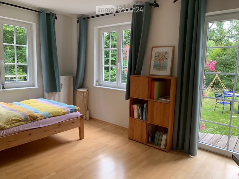 Bright bedroom with wooden flooring, large windows, a bed with a colorful quilt, a wooden bookshelf filled with books, and a framed picture on top of the shelf. A view of a garden is visible through a large window.