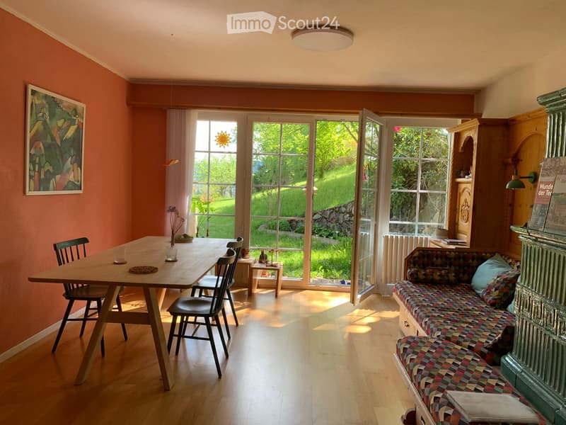 Bright living and dining area with wooden flooring, large windows, a colorful sofa, a wooden dining table with chairs, and a picture frame on the wall.