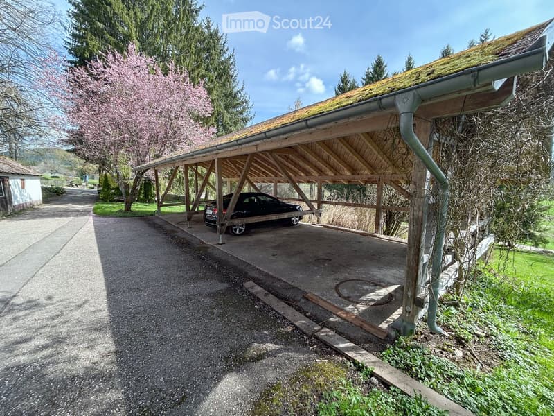 A wooden carport with a black car parked underneath, surrounded by greenery and a gravel driveway.
