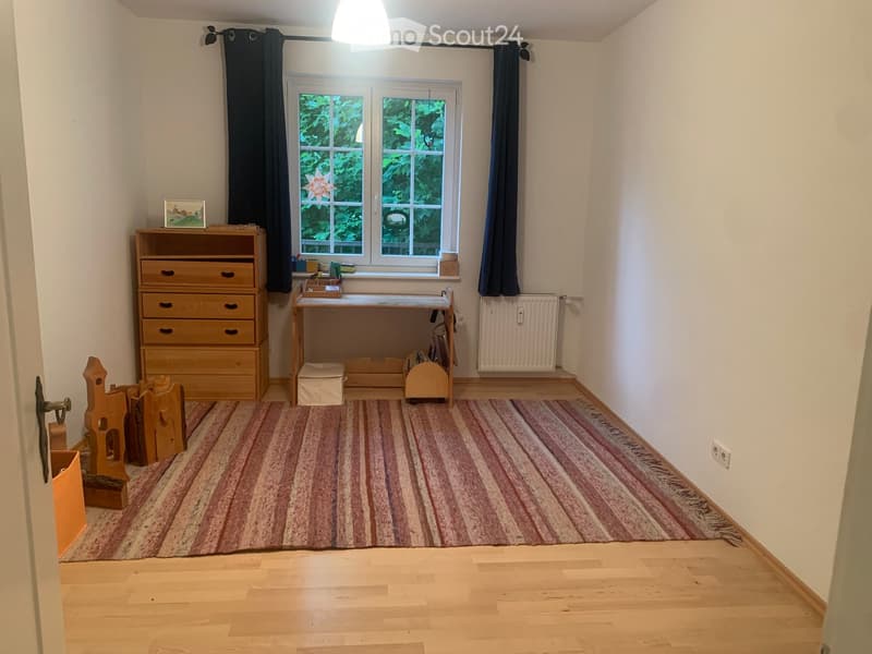 A small, well-lit room with wooden flooring, a large window with dark curtains, a striped rug, a wooden desk with a chair, a wooden drawer unit, a radiator, and a few toys.