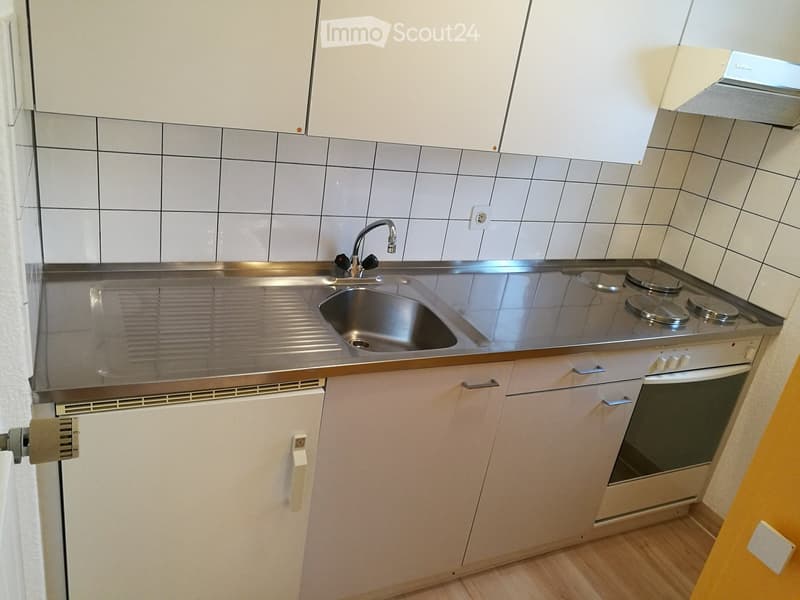 Modern kitchen with white cabinets, stainless steel sink, electric stove, and refrigerator. The walls are tiled in gray, and the floor is wooden.