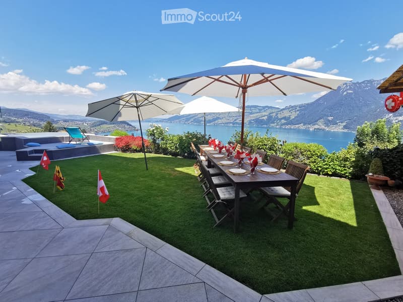 Outdoor patio with artificial grass, a dining table with chairs, large umbrellas, and a scenic view of a lake and mountains.