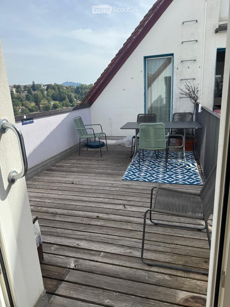 Wooden balcony with a table, chairs, and a bench, featuring a blue and white patterned rug and a scenic view of trees and hills.