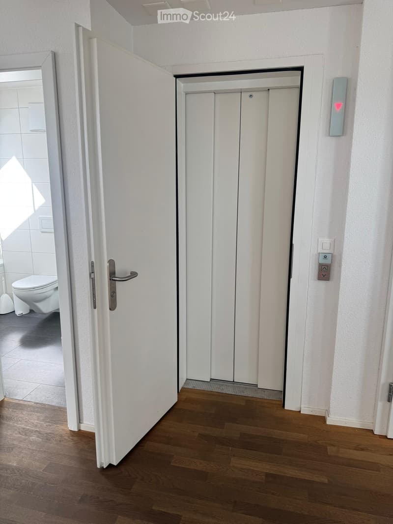 A hallway with wooden flooring, white walls, and an elevator door on the right. An open door on the left leads to a bathroom with a toilet and tiled walls.