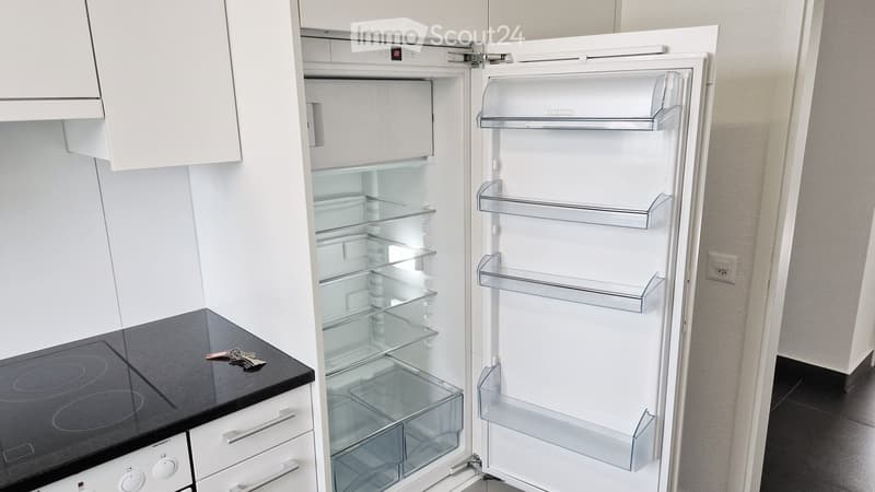 Modern kitchen with white cabinets, an open refrigerator, and an induction stove. The refrigerator is empty, and the stove has a set of keys placed on it.