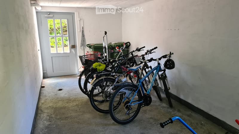 A narrow hallway with several bicycles parked inside, a door with a window, and a green trash bin in the background.