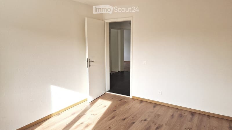 Empty room with light-colored walls and wooden flooring, featuring an open doorway leading to another room.