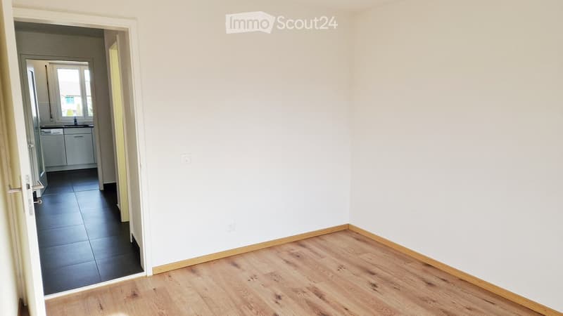 Empty room with wooden flooring and white walls, featuring a doorway leading to another room with tiled flooring and a sink.