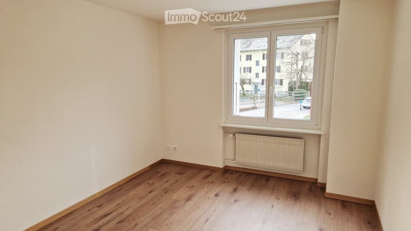 Empty room with wooden flooring, white walls, a window with white frame, and a radiator below the window.