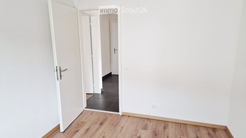 Empty room with light wooden flooring, white walls, and an open doorway leading to another room with dark flooring.