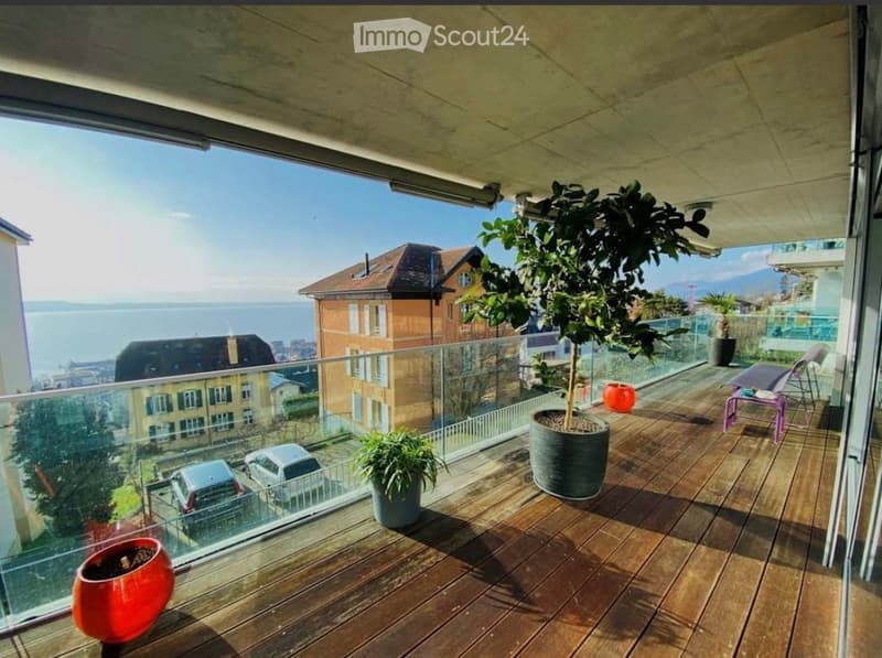 Balcony with wooden decking, glass railing, potted plants, and a view of the sea and surrounding buildings.