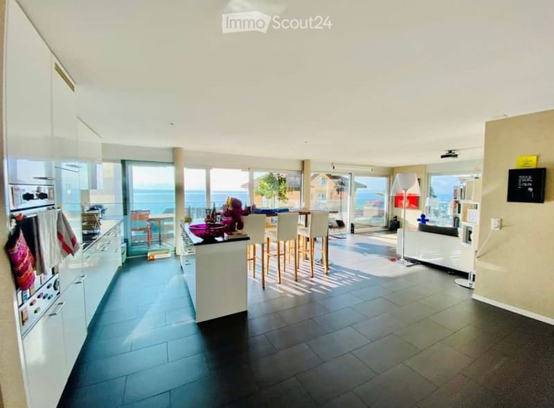 Modern kitchen and living area with large windows offering a sea view, equipped with a refrigerator, oven, kitchen island, bar stools, sofa, and floor lamp.