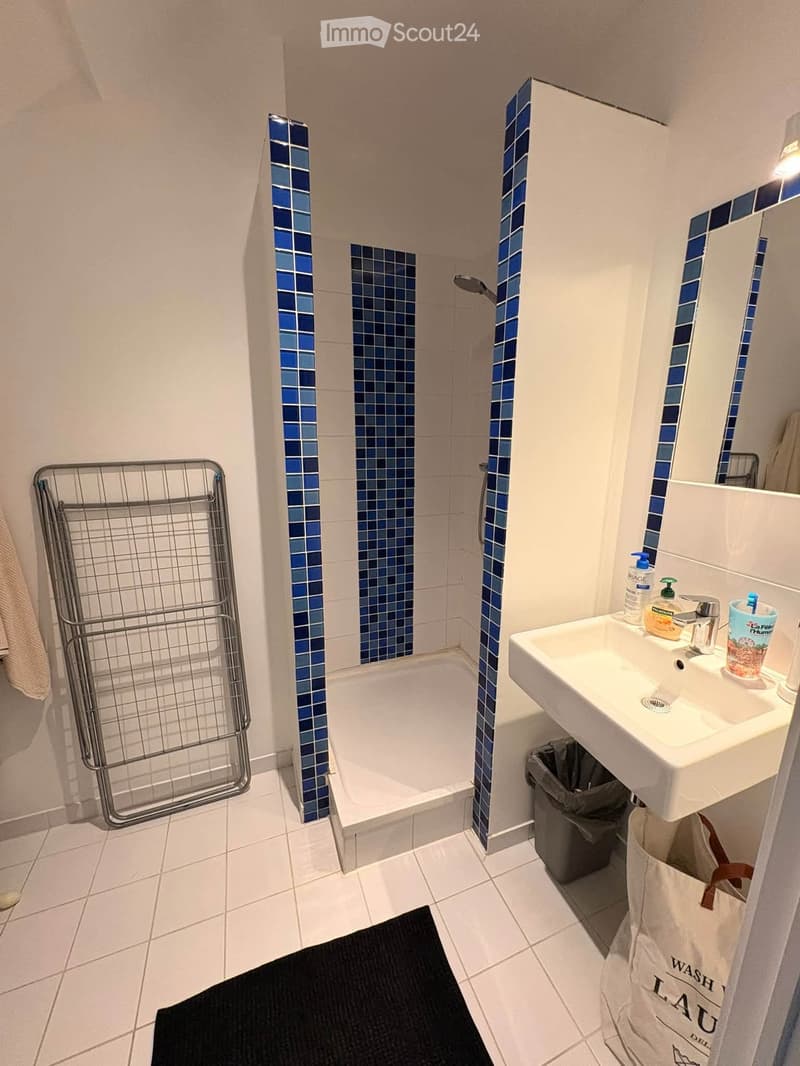 Modern bathroom with white tiles, blue mosaic accents, bathtub, sink, mirror, towel rack, and small trash bin.