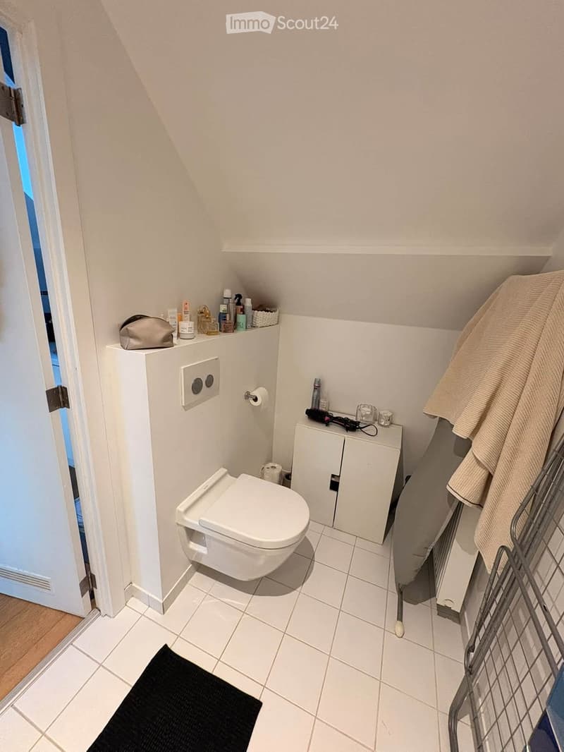 Compact bathroom with white tiled floor and walls, toilet, small cabinet, mirror, towel, and personal care items on shelves.