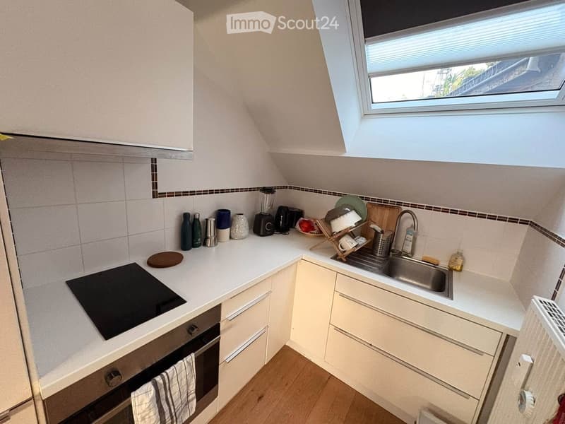 Compact kitchen with white cabinets, a black induction cooktop, and a stainless steel sink. The countertop holds various kitchen items including a blender, toaster, cups, and bottles.