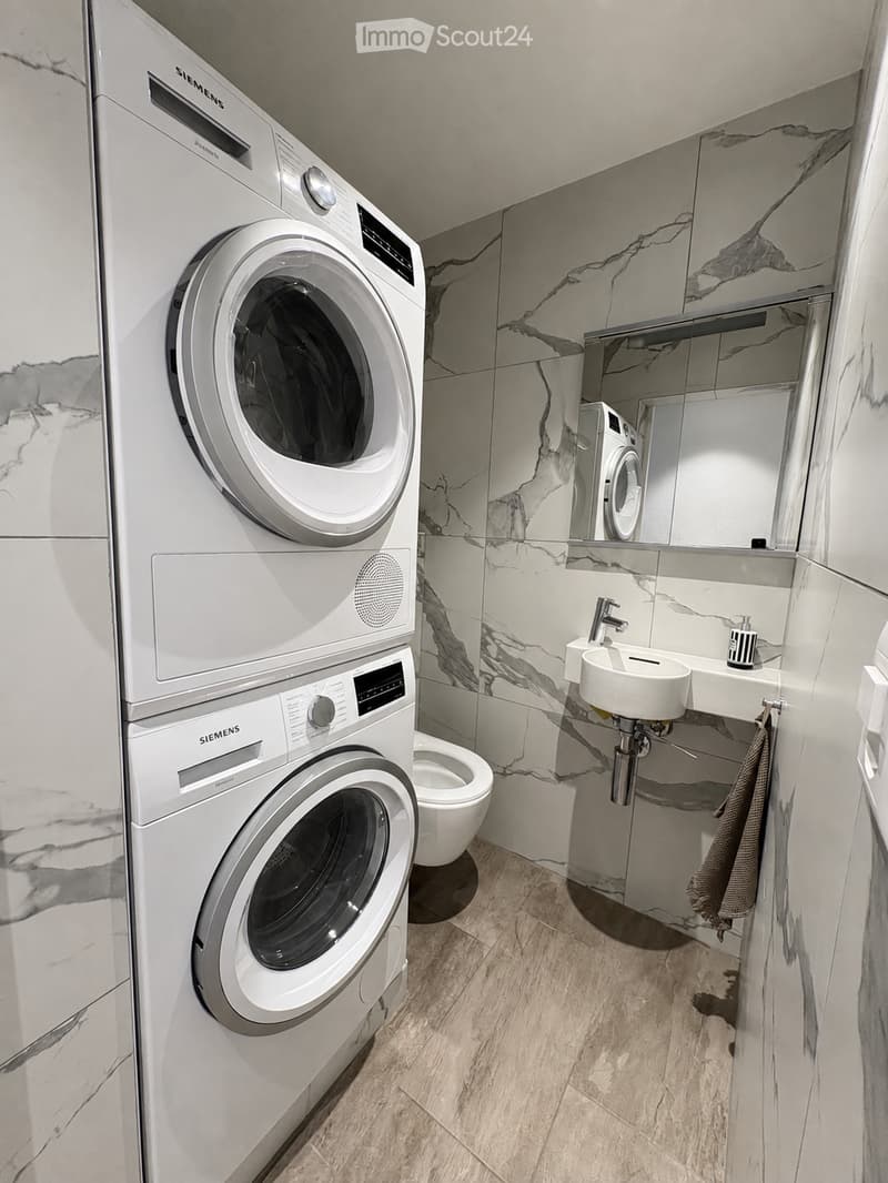 Modern bathroom with marble walls, a double sink, a toilet, and a stacked Siemens washing machine and dryer unit.