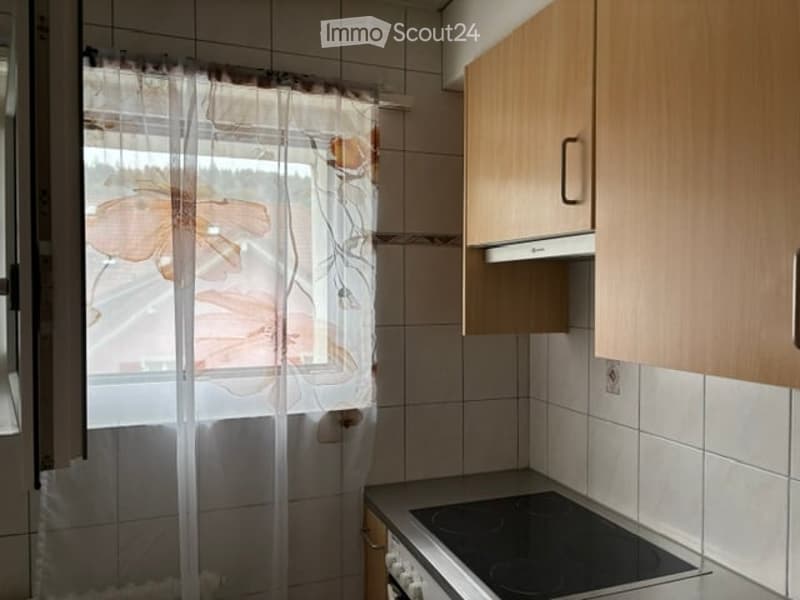 A small kitchen with tiled walls, wooden cabinets, and an electric stove. A window with a floral curtain allows natural light to enter.