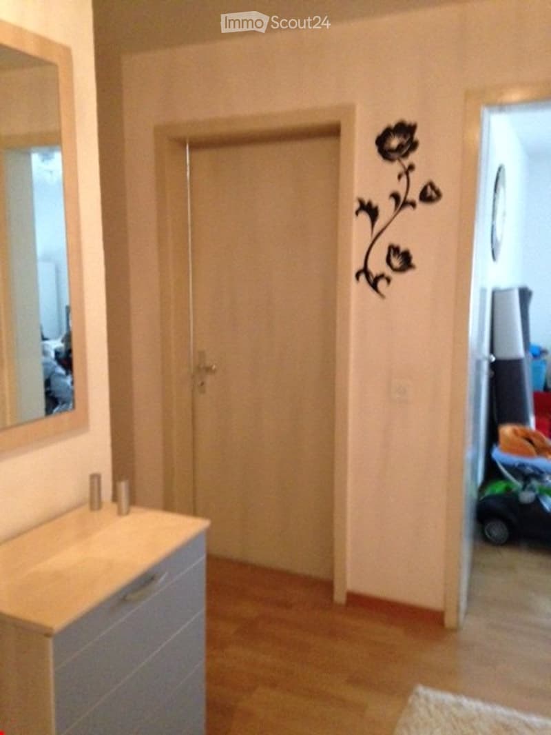 A narrow hallway with a wooden floor, a closed door, a wall-mounted mirror, and a decorative wall sticker featuring a flower design. A small cabinet with a drawer is visible on the left side.