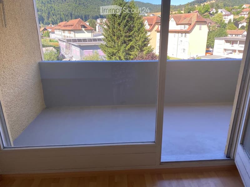 Balcony with a view of a town and surrounding greenery, featuring a glass railing and wooden flooring.